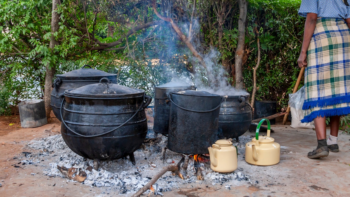 Photo depicts a woman cooking using four black cauldrons over an ashy and wood filled fire